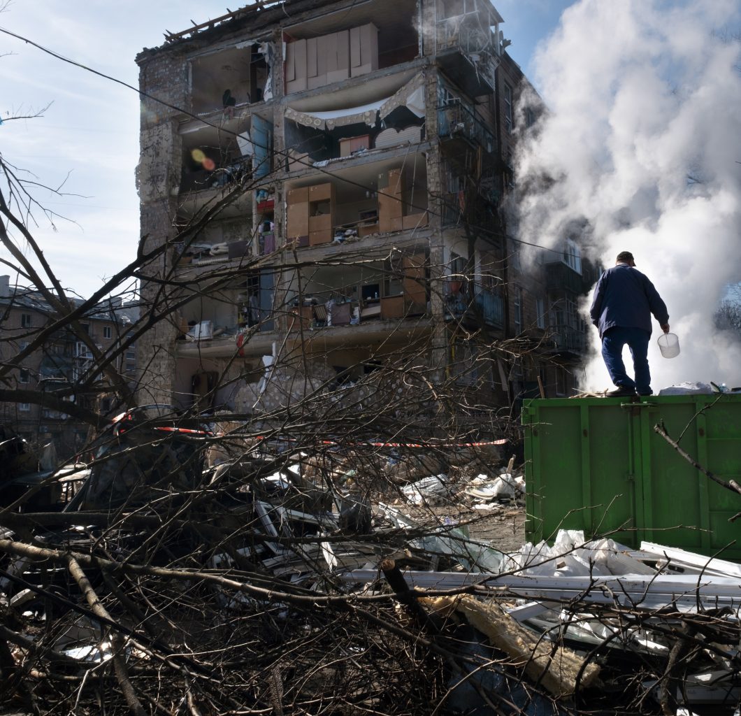 Landscape photo showing an area of Ukraine bombed with a build destroyed. A man faces with his back to the photo, standing on debris, looking at the building
