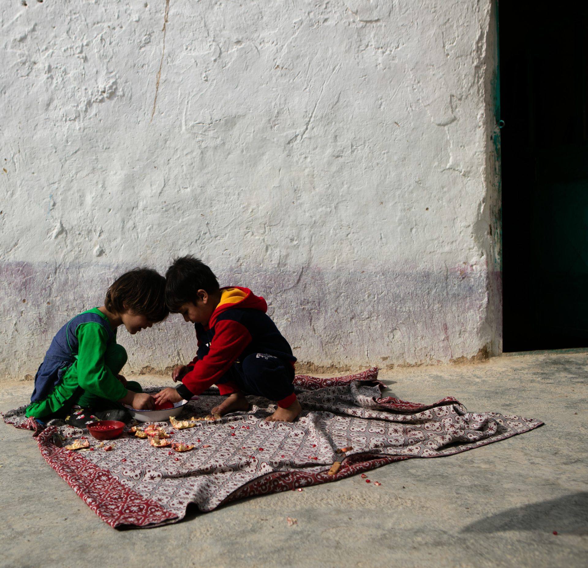 Two young children sat head to head on the floor outside their home in Afghanistan. They are sat on a patterned mat on hard floor.