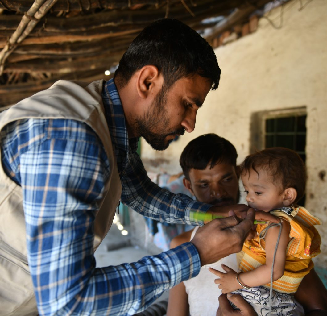 Action Against Hunger male staff member (on left) measuring a young boy's arm with MUAC band (on right). The young boy is being held by his father.