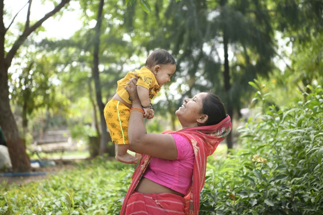 A smiling mother joyfully lifts her child outdoors. Action We support families like hers to access the nutrition and resources they need to survive.