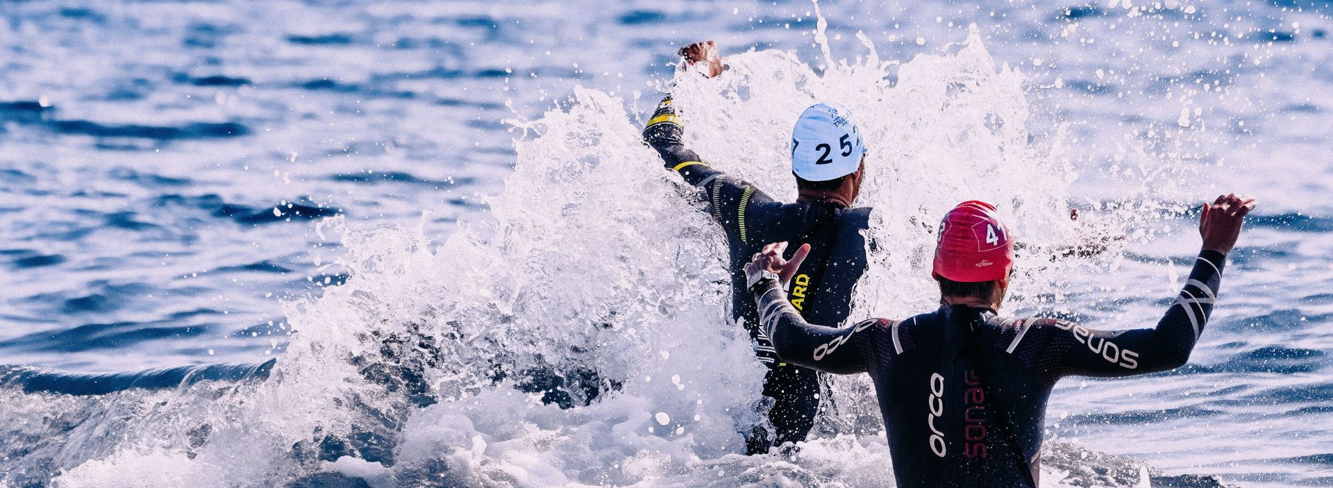 Two swimmers in wetsuits in lake among splashes.
